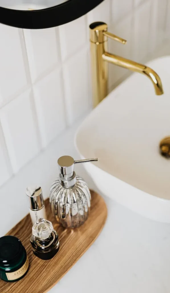 Minimalist bathroom sink with gold faucet and refillable glass soap dispenser on a wooden tray, styled for a non-toxic routine.